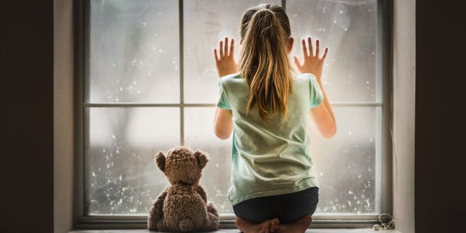 Mädchen mit Heimweh guckt aus dem Fenster mit Bär